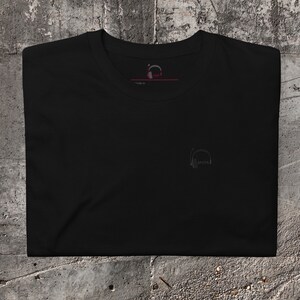 May include: Black t-shirt with a small, grey graphic of headphones with the word "SPOTS" printed inside. The shirt has a crew neck and short sleeves.