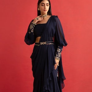 May include: A navy blue formal outfit. It includes a draped skirt, a matching top, and a sheer, long-sleeved top. The sleeves and belt are decorated with detailed embroidery. A gold-toned belt is at the waist, set against a red background.