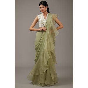 May include: A sage green ruffled saree with a white embroidered sleeveless blouse. The saree is draped in a modern style with multiple layers of ruffles.