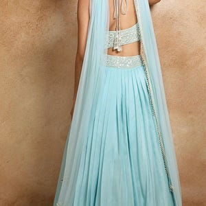 K&ouml;nnte beinhalten: Ein hellblaues Abendkleid mit einem plissierten Rock und einem passenden, durchsichtigen Schal. Das Oberteil und die Taille sind mit aufwendiger silberner Stickerei verziert. Der R&uuml;cken des Kleides ist offen, und der Schal ist &uuml;ber die Schultern drapiert.