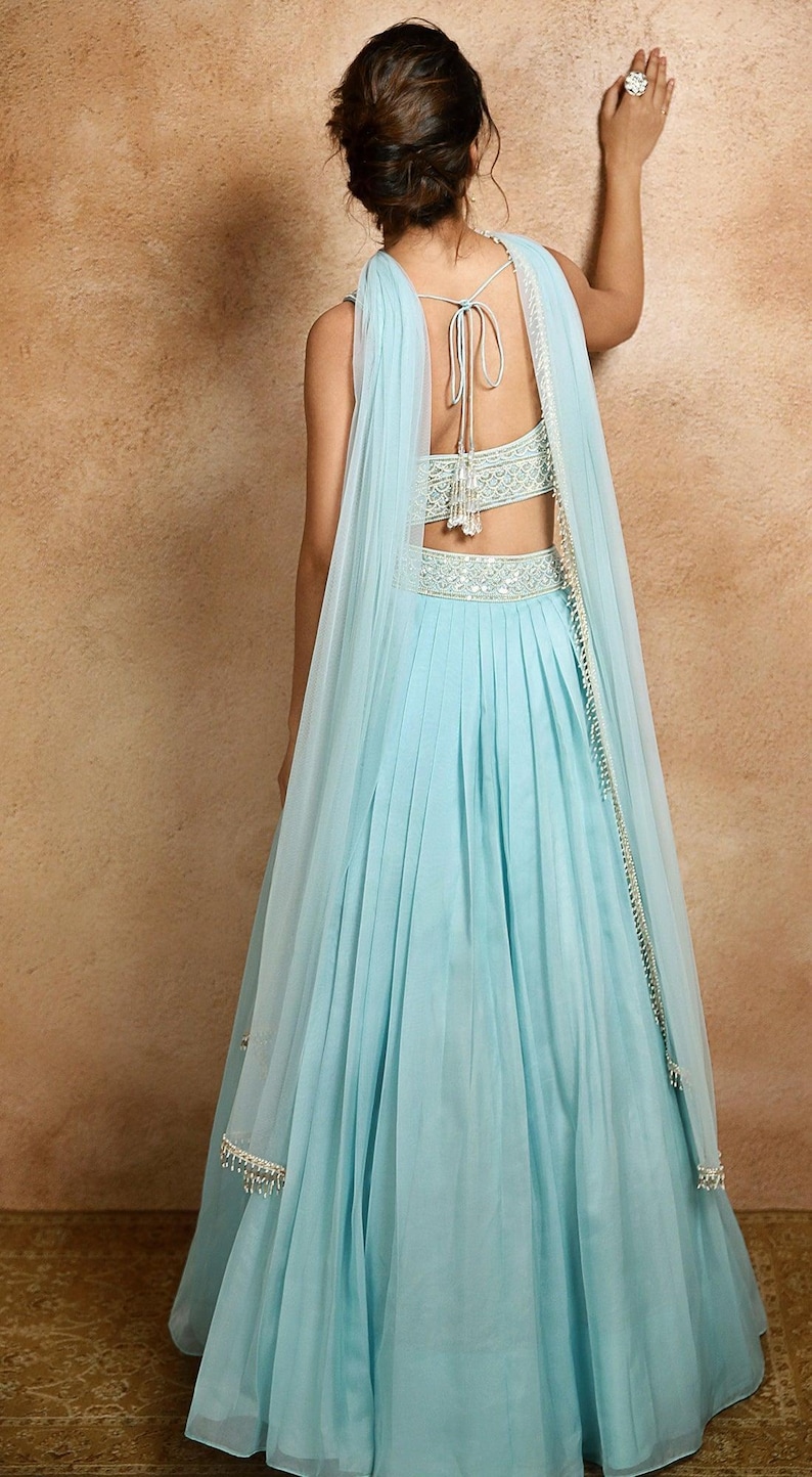K&ouml;nnte beinhalten: Ein hellblaues Abendkleid mit einem plissierten Rock und einem passenden, durchsichtigen Schal. Das Oberteil und die Taille sind mit aufwendiger silberner Stickerei verziert. Der R&uuml;cken des Kleides ist offen, und der Schal ist &uuml;ber die Schultern drapiert.