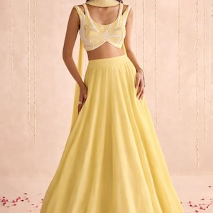 May include: A woman wearing a yellow lehenga choli, a traditional Indian outfit. The outfit includes a sleeveless crop top with white and yellow embroidery and a flowing skirt. The ensemble is accessorized with a matching scarf and earrings.