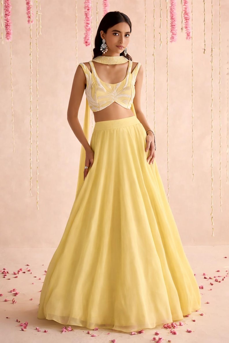 May include: A woman wearing a yellow lehenga choli, a traditional Indian outfit. The outfit includes a sleeveless crop top with white and yellow embroidery and a flowing skirt. The ensemble is accessorized with a matching scarf and earrings.