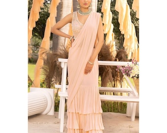 May include: A light peach-colored saree with a sleeveless, embellished top. The saree features a draped design and a tiered, ruffled skirt. The model is wearing a matching necklace and bracelet. The background includes decorative fabric and a white bench.