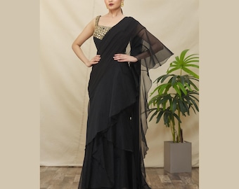 Puede incluir: Un vestido de noche negro con un corpiño dorado con lentejuelas y una falda en capas. El vestido presenta un diseño de un solo hombro con una superposición de gasa fluida. La modelo lleva pendientes dorados.