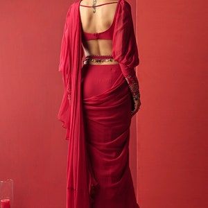 May include: A red saree with a low back and long sleeves. The saree is draped in a way that creates a flowing, elegant look. The saree is paired with a matching red blouse.