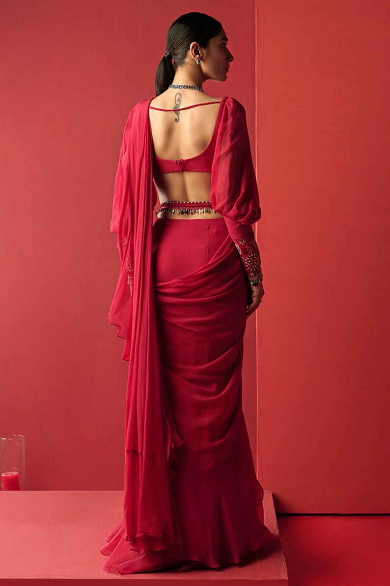 May include: A red saree with a low back and long sleeves. The saree is draped in a way that creates a flowing, elegant look. The saree is paired with a matching red blouse.