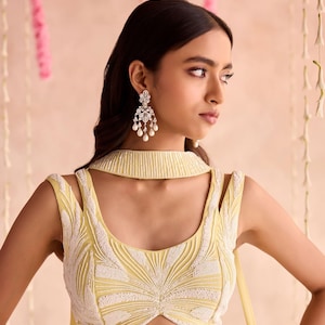 May include: A light yellow sleeveless top with white beaded embroidery. The top has a high collar and cut-out shoulder straps. The model is wearing silver earrings with dangling pearls. The background is a soft pink.