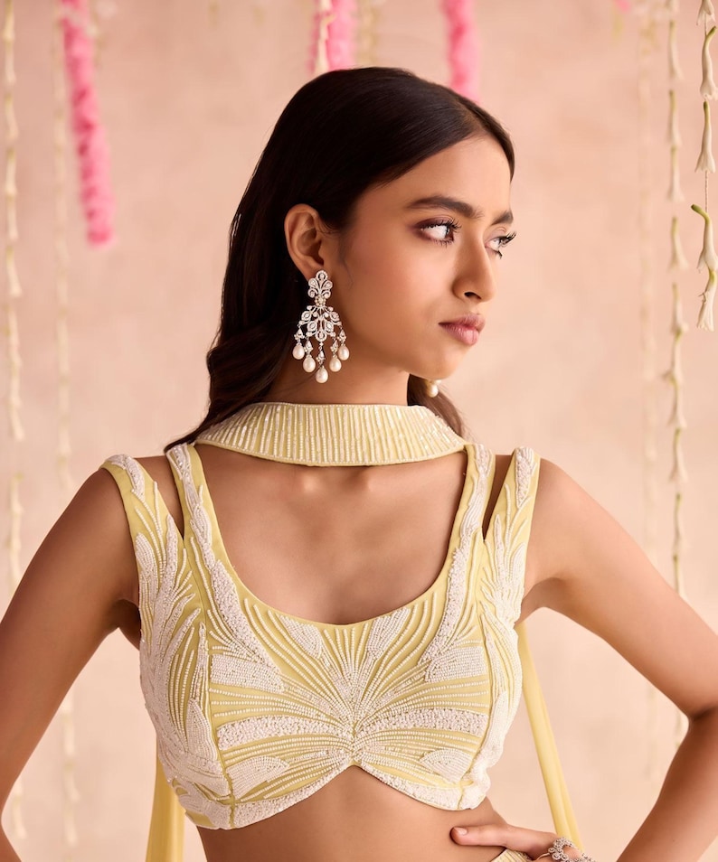 May include: A light yellow sleeveless top with white beaded embroidery. The top has a high collar and cut-out shoulder straps. The model is wearing silver earrings with dangling pearls. The background is a soft pink.