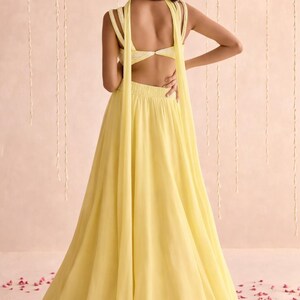 May include: A back view of a yellow formal dress with a flowing skirt and open back. The dress has a halter-style top with straps that drape down the back. The fabric appears lightweight and airy.