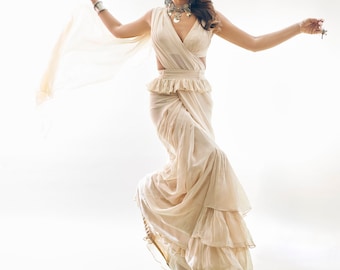 May include: A woman wearing a white, ruffled saree with a plunging neckline and silver jewelry. She is smiling and holding a small object in her right hand.