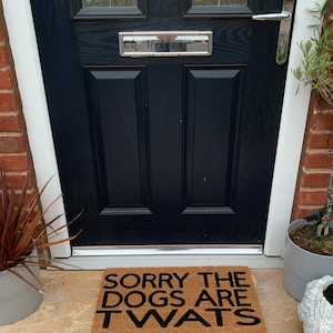 May include: A coir doormat with the phrase "SORRY THE DOGS ARE TWATS" in black lettering. The mat sits in front of a dark blue door with a silver handle and a sign that says "Oh Hello!".
