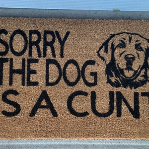 May include: A brown coir doormat with the text "SORRY THE DOG IS A CUNT" in black lettering. A black silhouette of a golden retriever's head is centered above the text.