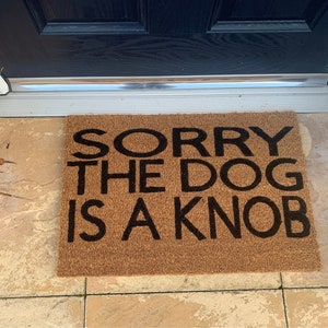 May include: A brown coir doormat with the text "SORRY THE DOG IS A KNOB" in black lettering.