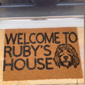 May include: A brown coir doormat with the text "WELCOME TO RUBY'S HOUSE" in black lettering. A black silhouette of a dog's head is to the right of the text.