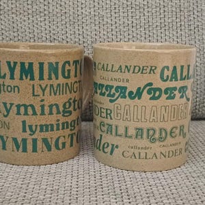 May include: Two ceramic mugs with a speckled tan finish. The left mug features the word Lymington printed in various green fonts and sizes. The right mug features the word Callander printed in various green fonts and sizes across the surface.