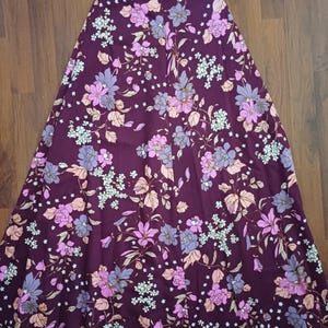 May include: A long, flowing skirt in a deep burgundy hue, patterned with pink, purple, and light green flowers, and beige leaves. The skirt is made from a soft fabric. The floral design is spread across the skirt.