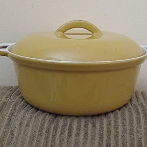 May include: A yellow cast iron casserole dish with a matching lid. The pot has two white-rimmed handles and a loop handle on the lid. The pot is sitting on a brown, textured surface.