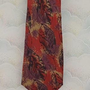 Puede incluir: Una corbata roja con un patrón repetido de hojas grandes y estilizadas en tonos burdeos, morados y beige. La corbata tiene una forma clásica y está hecha de una tela lisa.
