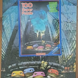 May include: A 100-piece Teenage Mutant Ninja Turtles jigsaw puzzle featuring a city skyline and the four turtles emerging from a manhole cover. The box displays the title 100 Super Puzzle, the Waddingtons logo, and includes a poster measuring 68 x 48 cm.