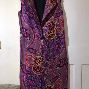 May include: A long purple sleeveless vest with a floral pattern in shades of pink, orange, and gold. The vest has a collar and a button closure.
