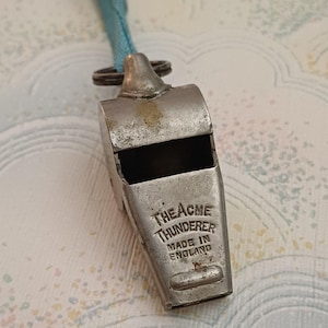 May include: A vintage metal whistle with a light blue lanyard. The whistle is marked "THE ACME THUNDERER MADE IN ENGLAND". The whistle has a rectangular body with a rounded top and a small ring for the lanyard.