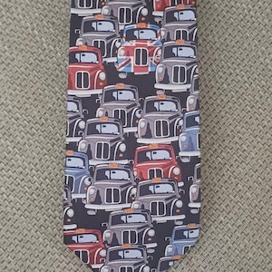 Puede incluir: Una corbata negra con un patrón repetitivo de taxis londinenses en varios colores, incluyendo rojo, azul y gris. Cada taxi tiene un pequeño letrero naranja en el techo. Un taxi en el centro muestra un diseño de la bandera Union Jack en el capó.
