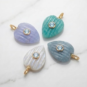 May include: Four heart-shaped pendants in light blue, teal, white, and grey, each featuring a ribbed texture and a central round light blue gemstone set in a gold-toned bezel. Each pendant includes a small gold-toned beaded bail at the top.