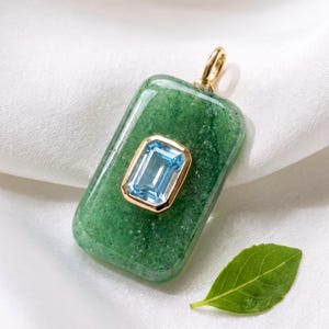 May include: A rectangular green aventurine pendant featuring a central emerald-cut light blue gemstone set in a gold-toned bezel. The pendant includes a gold-toned bail at the top for attachment to a chain, showcasing a smooth, polished surface.