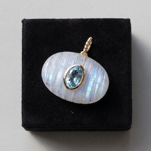 May include: An oval-shaped pendant featuring a translucent, ribbed moonstone with a blue iridescent sheen. A faceted, oval-cut light blue gemstone is bezel-set in gold at the center, topped with a small, textured gold bail for attachment.