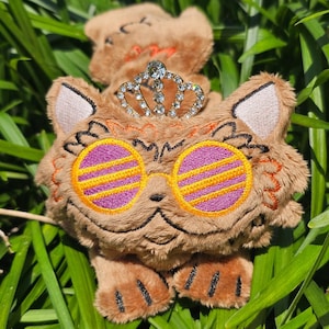 May include: A brown plush cat toy featuring embroidered details, including large round glasses with yellow frames and purple lenses. The cat wears a silver-toned metal crown adorned with clear rhinestones on its head. The plush has stitched paws.