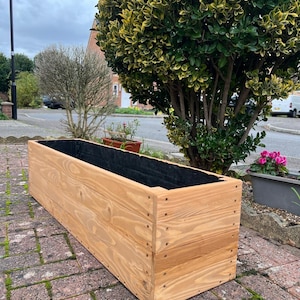 May include: A large, rectangular wooden planter box made from light-coloured wood. The box has a black interior lining and is held together with screws. The planter sits on a brick surface, with a cloudy sky and greenery in the background.
