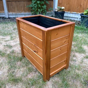 May include: A wooden rectangular planter, constructed from horizontal wooden slats, with a natural finish. It features a black liner and is positioned on grass. Ideal for gardens, the planter is approximately 60cm long.