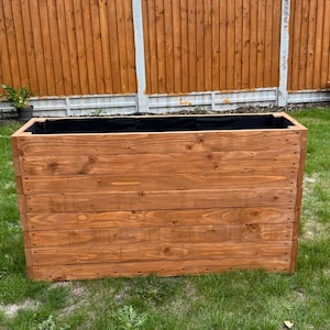 May include: A rectangular wooden planter box made from stained timber. The box is constructed with horizontal planks and has a black interior lining. It sits on grass, with a wooden fence in the background.