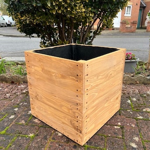 May include: A large, square wooden planter box with a natural wood finish. The box is constructed from horizontal planks and has a dark interior. The planter is sitting on a brick surface, with a tree and houses in the background.