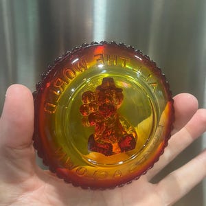 May include: A vintage, round, amber-coloured glass dish with a clown figure in the centre. The rim has a beaded design, and the words "A VICTORIA" and "THE MOST" are visible around the edge. The dish is held in a hand.