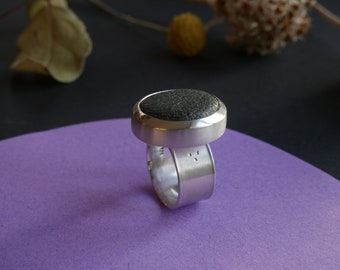 May include: A silver ring with a black stone set in a round bezel. The ring is on a purple surface.