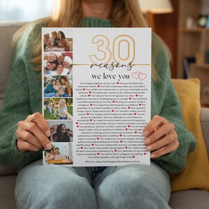 May include: A white card with gold lettering that reads "30 reasons we love you" with two red hearts. The card has a collage of photos of people smiling and laughing. The card is being held by a person's hands.