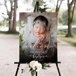 May include: A black welcome sign with white text that reads "Welcome to Monica's Baptism 08.12.2024". A photo of a baby girl wearing a headband is in the background.