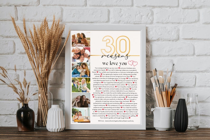 May include: A white framed print with a gold number 30 and the words "30 reasons we love you" in a gold font. The print features a collage of photos of people and a list of reasons why they are loved. The print is on a wooden table with a vase of wheat stalks and a white vase with paintbrushes.
