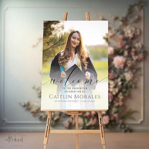 May include: A graduation welcome sign on a wooden easel. The sign features a photo of a graduate in a cap and gown, with the word "welcome" in cursive. Text below reads "To the Graduation Celebration of Caitlin Morales, Signature University, Class of 202X."