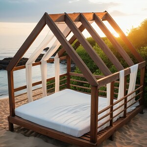 Könnte beinhalten: Ein Holz-Baldachinbett mit weißen Vorhängen und einer weißen Matratze. Das Bett steht auf einem Sandstrand mit üppigem Grün im Hintergrund.