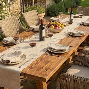 May include: A wooden outdoor dining table set for a meal, surrounded by woven wicker chairs. The table has a cream-coloured runner, plates, wine glasses, and a bowl of fruit. Wine bottles are also present. The table is approximately 240 cm long.