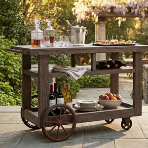 May include: A wooden outdoor bar cart with a rustic design. It features shelves for bottles, glasses, and a serving tray. The cart has large spoked wheels and is filled with drinks, snacks, and fruit. The cart is set on a stone patio.