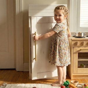 May include: A young child in a floral dress opens a white toy refrigerator with a gold handle. A wooden toy kitchen set with pots and pans is visible. The scene is set in a well-lit room with a wooden floor and a rug.