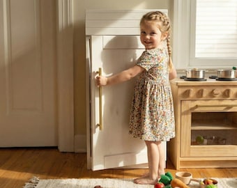 May include: A young child in a floral dress opens a white toy refrigerator with a gold handle. A wooden toy kitchen set with pots and pans is visible. The scene is set in a well-lit room with a wooden floor and a rug.