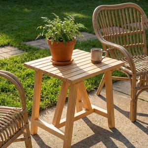 May include: A small, light-colored wooden side table with a square top and angled legs. A potted plant and a mug sit on the table. Two wicker chairs are in the background, with green grass visible.