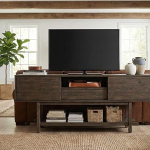 May include: A living room scene featuring a large dark wood entertainment center with a TV. Two brown leather armchairs with a blanket are on either side. A fireplace and large windows are in the background. Decorative plants and baskets add to the decor.