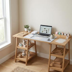 Puede incluir: Un escritorio de madera clara con una computadora portátil, una impresora y varios suministros de artesanía. El escritorio tiene dos estantes con rollos de cinta decorativa y cuerda. Una taza, tijeras y pequeñas plantas también están en el escritorio.