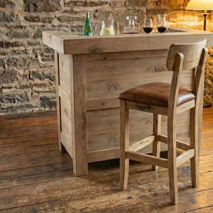 Op de afbeelding: Een rustieke houten bar met bijpassende barkruk. De bar is gemaakt van lichtgekleurd hout en heeft een aanrecht met glaswerk en flessen. De kruk heeft een bruine leren zitting. De kamer heeft een stenen muur en een houten vloer.